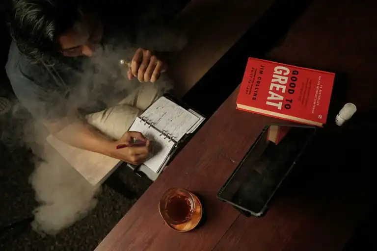 成年使用者在桌前書寫筆記並使用電子煙，桌上放有平板與書籍