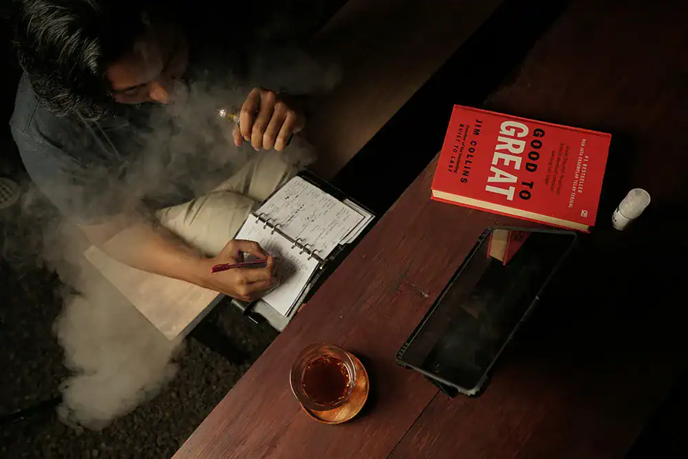成年使用者在桌前書寫筆記並使用電子煙，桌上放有平板與書籍